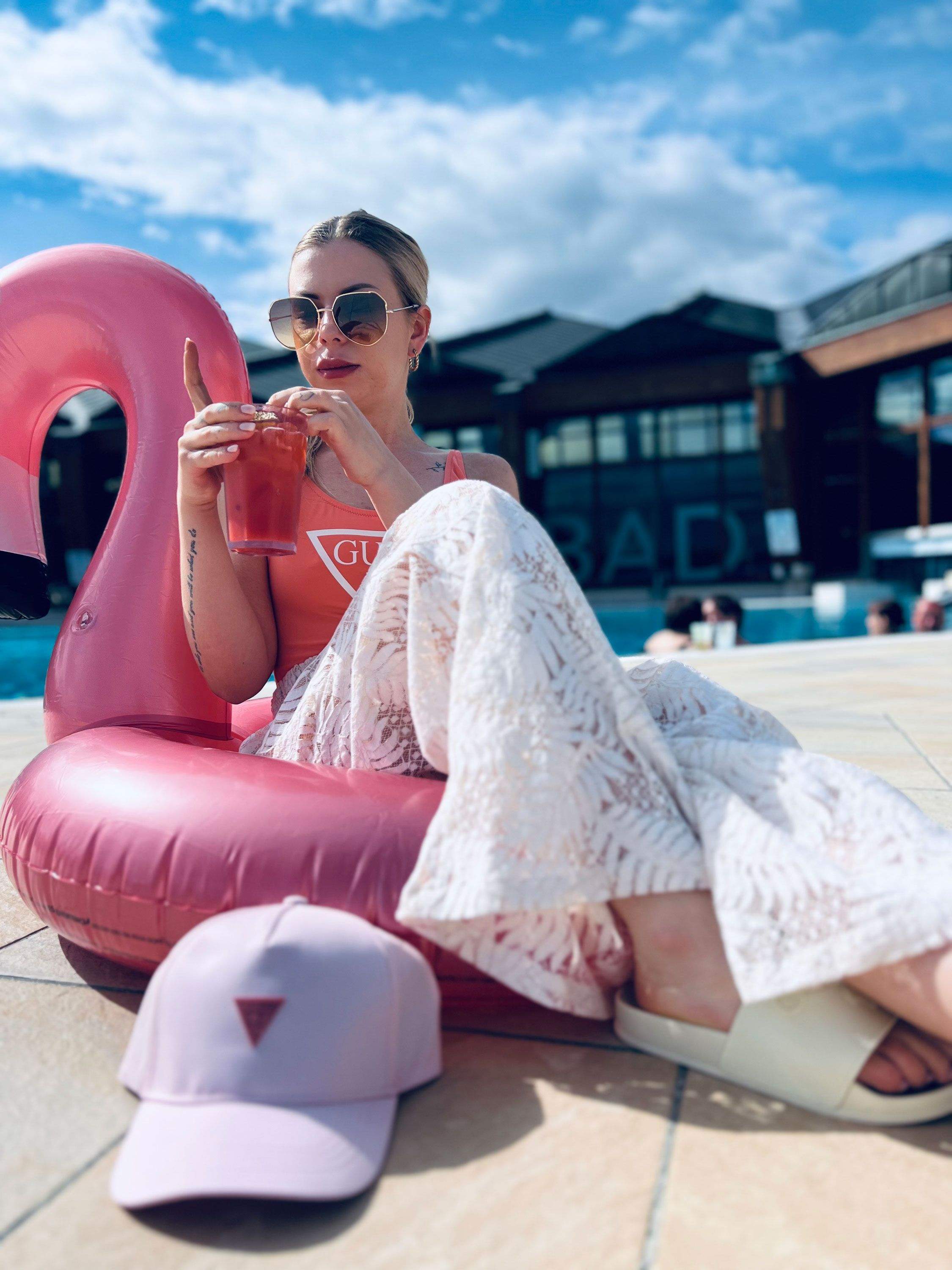 Person sitzt auf einem aufblasbaren pinkfarbenen Flamingo, trägt weiße Hose mit Muster und weiße Sandalen, hält ein Getränk in der Hand, vor ihr liegt eine hellrosa Kappe mit rotem Dreieck-Logo.