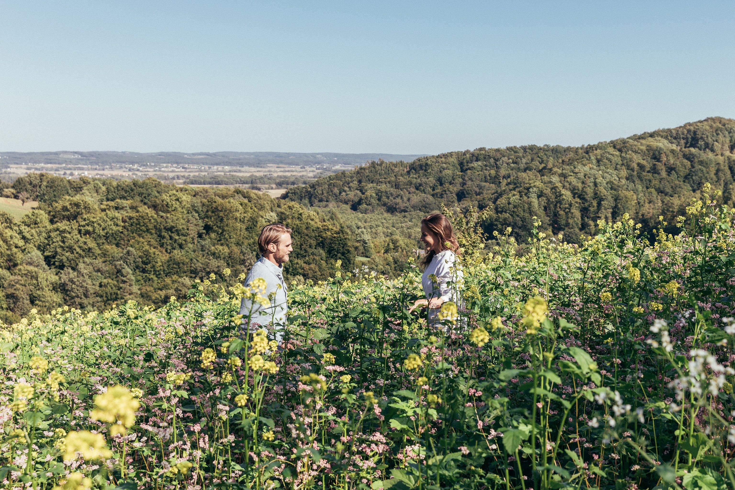 Couple standing in the meadow and the landscape around Bad Loipersdorf can be seen in the background.