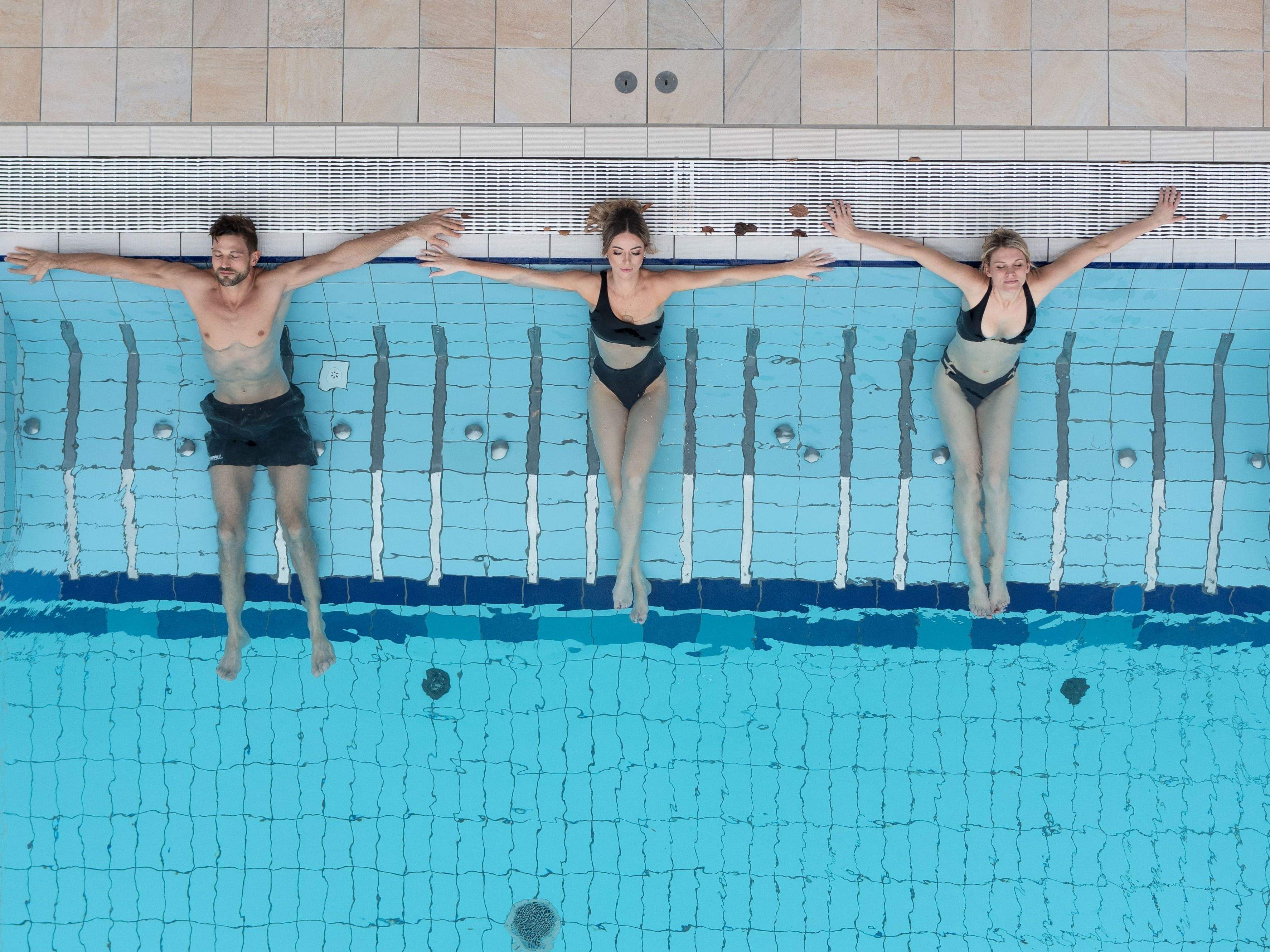 Drei Personen liegen entspannt am Beckenrand eines Schwimmbeckens mit ausgebreiteten Armen, zwei Frauen in Badeanzügen und ein Mann in Badeshorts.