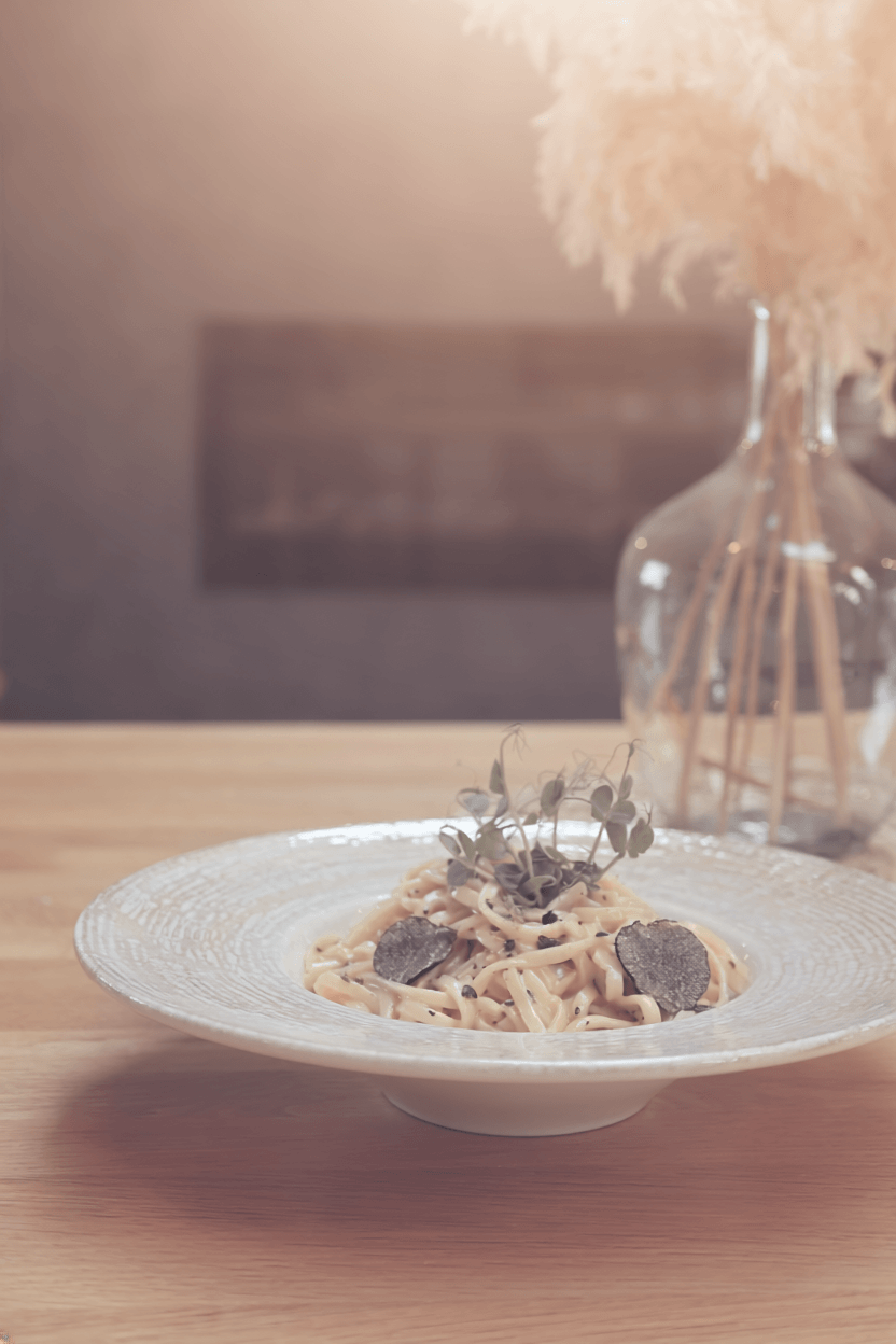 Plate of pasta garnished with truffle slices and fresh herbs on a wooden table in front of a vase of dried flowers.