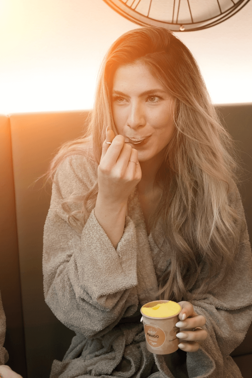 Frau mit langem, gewelltem Haar in Bademantel sitzt auf Sofa und hält Becher mit Eiscreme in der Hand