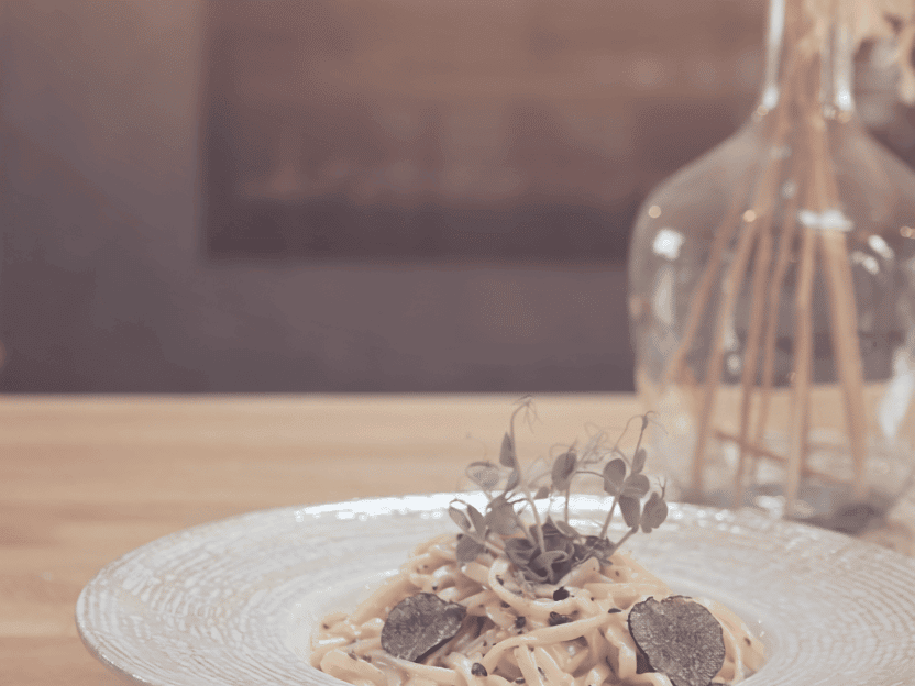 Teller mit Pasta, garniert mit Trüffelscheiben und frischen Kräutern, auf einem Holztisch vor einer Vase mit getrockneten Blumen.