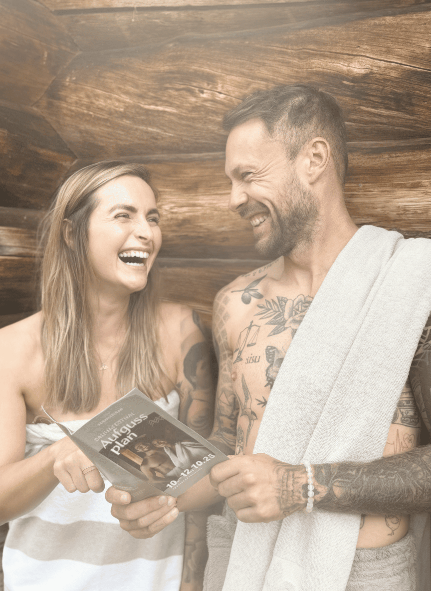 Zwei tätowierte Personen in Saunatüchern lesen gemeinsam einen Plan in einer Holzsauna